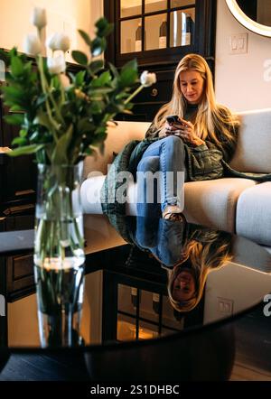 Die junge Frau sitzt auf der Couch und sucht auf dem Handy Stockfoto