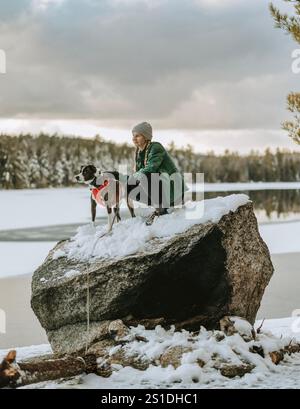 Frau sitzt auf schneebedeckten Felsen am See mit ihrem Hund in Maine Stockfoto