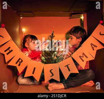 Zwei Kinder in festlichen Pullover sitzen in einem Karton mit Lichterketten und einem kleinen Weihnachtsbaum. Bruder und Schwester machen Ornament Stockfoto