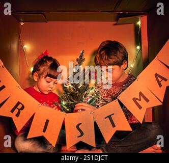 Zwei Kinder in festlichen Pullover schmücken einen kleinen Weihnachtsbaum in einem beleuchteten Karton. Ein Banner mit der Schreibweise WEIHNACHTEN hängt an der Schachtel Stockfoto