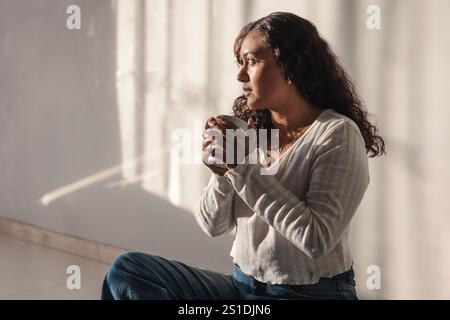 Eine junge Frau, die eine Tasse Kaffee hält und durch ein Hausfenster schaut. Stockfoto