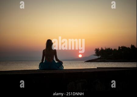 Eine Frau in blauem Kleid sitzt auf einem Felsvorsprung und blickt auf einen ruhigen Sonnenuntergang oder Sonnenaufgang über ruhigem Wasser mit Silhouetten von Bäumen und Bergen im Hintergrund Stockfoto