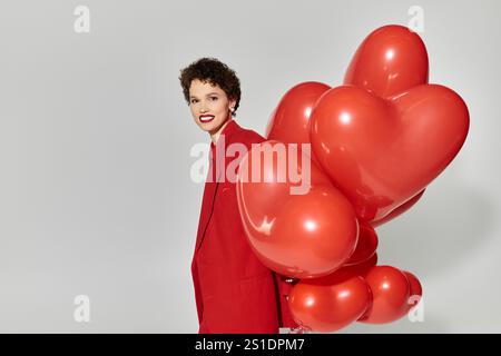 Eine schöne junge Frau, die hell lächelt, während sie einen Haufen roter Herzballons hält. Stockfoto