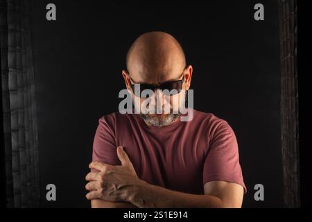 Ein kahler bärtiger Mann trägt eine Sonnenbrille mit überkreuzten Armen. Isoliert auf dunklem rauchigen Hintergrund. Person aus Südamerika. Stockfoto