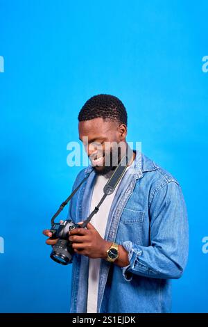 Der junge Fotograf lächelt glücklich, während er Fotos auf seiner Kamera vor einem leuchtend blauen Hintergrund in einer Studioaufnahme ansieht Stockfoto