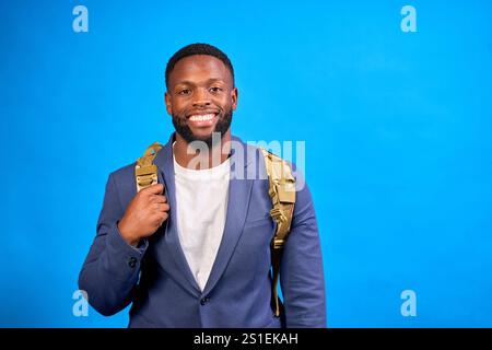 Studio-Porträt eines fröhlichen jungen Geschäftsmannes mit Rucksack, das Selbstvertrauen und Stil vor einem leuchtend blauen Hintergrund ausstrahlt Stockfoto