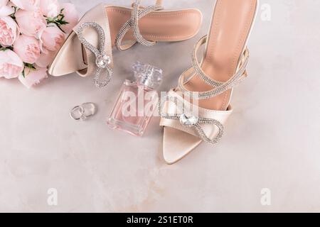 Ein Paar beige Satin-Schuhe mit einer Strassschleife auf beigefarbenem Hintergrund mit einem Strauß Pfingstrosen. Flasche pinkfarbenes Parfüm. Klingeln Stockfoto