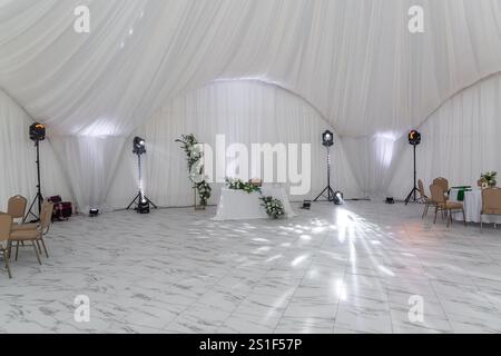 Weingläser, silbernes Geschirr und grüne Servietten auf runden Tischen mit weißer Tischdecke. Catering-Konzept. Innenraum des Zeltes für Hochzeitsessen, Stockfoto