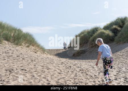 Harlech Wales - 1. August 2024; Frau in blumigen Hosen, die zwischen Dünen auf ein Paar mit Hunden zugeht, die in Richtung Harlech Beach in Nordwales kommen. Stockfoto