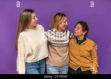 Enkelin, Mutter und Großmutter umarmen sich gegenseitig, feiern Familienbande und weibliche Ermächtigung vor einem leuchtend violetten Hintergrund und schaffen eine che Stockfoto