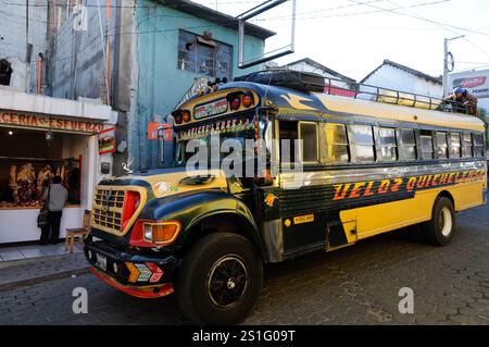 Hühnerbus-Öffentlicher Verkehr Chichicastenango Guatemala | CHICHICASTENANGO, Guatemala — Ein farbenfroher Hühnerbus schlängelt sich durch eine der belebten Straßen im nördlichen Ende der Innenstadt von Chichicastenango, einer hochland-indigenen Maya-Stadt, die für ihre lebendige Kultur bekannt ist. Hühnerbusse, umfunktionierte und farbenfroh dekorierte ehemalige nordamerikanische Schulbusse, dienen als primärer öffentlicher Verkehr in ganz Guatemala und verbinden Städte und Dörfer über das bergige Gelände hinweg. Chichicastenango liegt auf einer Höhe von fast 6.500 Fuß (1.980 Meter) im Departamento Quiché, etwa 90 Meilen (1 Stockfoto