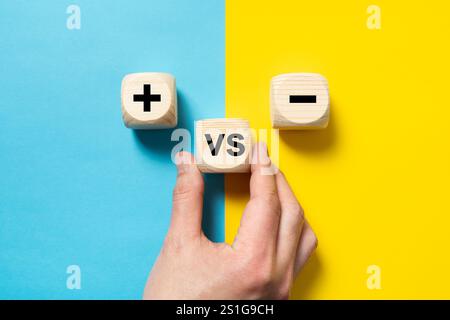 Händisch platzieren eines Holzblocks im Vergleich zum Plus- und Minuszeichen auf blauem und gelbem Hintergrund, Konzept der Gegensätze, vor- und Nachteile, flache Komposition Stockfoto