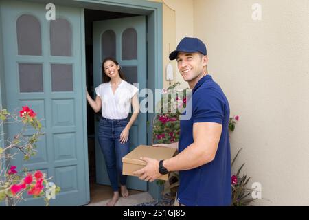 Positiver, gutaussehender Lieferer in blauer Uniform mit Pappschachtel Stockfoto