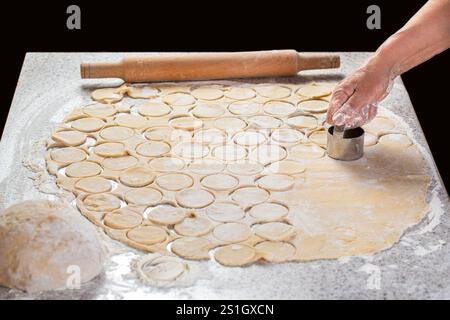 Zubereitung des Teigs zum Backen, großes Stück des Teigs, Koch schneidet kleine Kreise auf ihm in eiserner Form aus. Männliche Ravioli mit Rollen Stockfoto