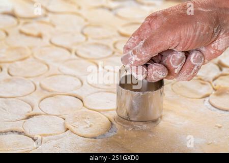 Zubereitung des Teigs zum Backen, ein großes Stück des Teigs liegt auf dem Tisch und der Koch schneidet kleine Kreise mit eiserner Form aus. Schneiden Stockfoto