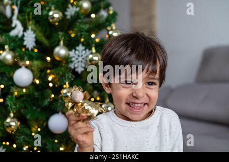 Ein kleiner Junge hält ein goldenes Ornament in der Hand, lächelt und schaut in die Kamera. Die Szene spielt in einem Wohnzimmer mit einem Weihnachtsbaum i Stockfoto