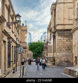 Baku, Aserbaidschan - 5. Mai 2024: Besucher schlendern durch die historischen engen Gassen von Icherisheher, umgeben von alter Architektur, während sich die modernen Flammentürme in der Ferne erheben Stockfoto