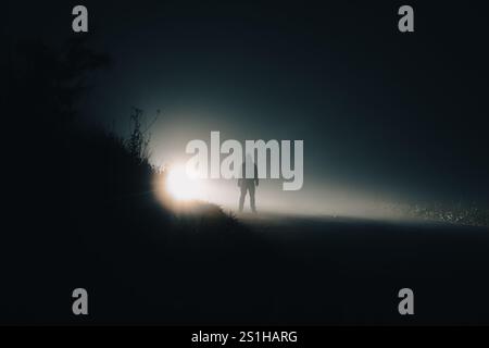 Eine gruselige Kapuzenfigur. Silhouette auf einer Landstraße. Auf einer geheimnisvollen nebligen Winterstraße bei Nacht Stockfoto