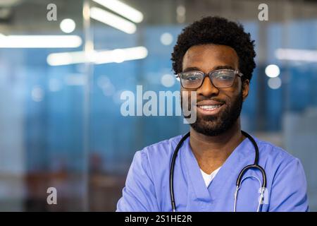 Nahporträt eines jungen Arztes, der Mann lächelt und in die Kamera schaut. Der Arzt trägt einen blauen medizinischen Mantel und ein Stethoskop und arbeitet in der Klinik. Stockfoto