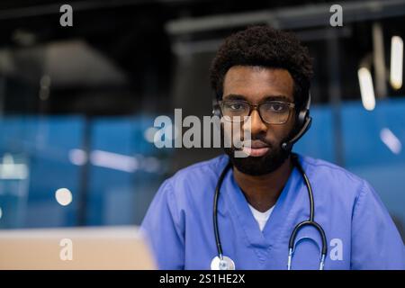 Junger Arzt mit Headset in die Kamera schauen, Nahaufnahme des Arztes in der Praxis. Mann in blauem medizinischem Mantel und Stethoskop, der aus der Ferne arbeitet, Klinikpatienten berät und mit Rat hilft. Stockfoto