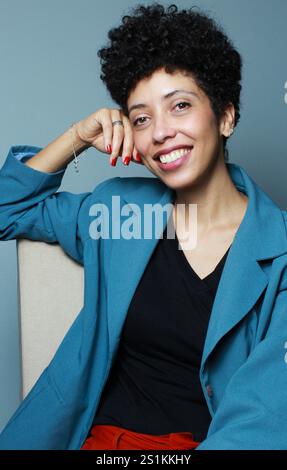 Erfolgreiche Geschäftsfrau sitzt entspannt auf einem Stuhl und lächelt in die Kamera. afroamerikanisches weibliches Model mit kurzen lockigen Haaren trägt Bürokleidung. Stockfoto