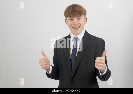 Glücklicher, sechzehnjähriger Teenager, der einen Anzug trägt, der die Daumen hoch hält, Studioaufnahme vor weißem Hintergrund Stockfoto