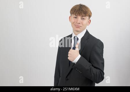 Glücklicher, sechzehnjähriger Teenager, der einen Anzug trägt, der die Daumen hoch hält, Studioaufnahme vor weißem Hintergrund Stockfoto