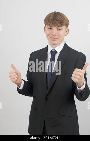 Glücklicher, sechzehnjähriger Teenager, der einen Anzug trägt, der die Daumen hoch hält, Studioaufnahme vor weißem Hintergrund Stockfoto