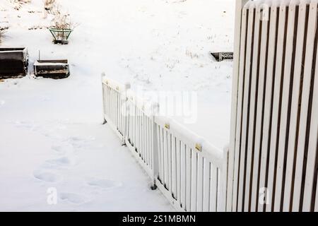 Schneebedeckter weißer Holzzaun mit sichtbaren Fußabdrücken im Neuschnee, Hochbeeten und Gestrüpp. Stockfoto