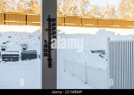 Das Außenthermometer auf Holzpfosten zeigt die Temperatur unter Null im schneebedeckten Hinterhof mit erhöhten Gartenbeeten und weißem Zaun an. Stockfoto