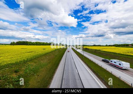 Gerade Autobahn, die durch gelbe Rapsfelder unter teilweise bewölktem Himmel führt, mit Autos und Wohnwagen. Stockfoto