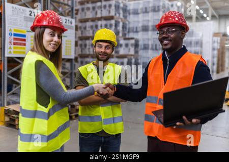 Drei Lagerarbeiter, die Sicherheitswesten und Helme tragen, arbeiten effektiv zusammen. Ein Mitarbeiter hält einen Laptop in der Hand, während die anderen die Hände schütteln Stockfoto