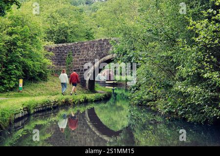 Ein Paar spaziert entlang des Caldon Canal in der Nähe der alten Brücke und nähert sich einem Schmalboot mit Reflexionen im Wasser an sonnigen Tagen - in der Nähe von Wetley Rocks, Staffordshire Stockfoto