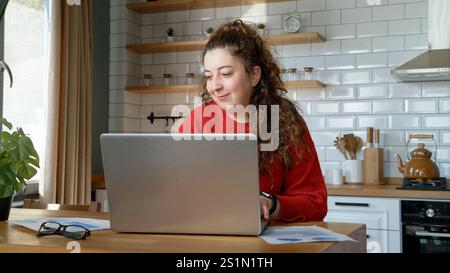 Eine fröhliche Frau in einer modernen Küche nutzt ihren Laptop und genießt einen entspannten Moment, während sie von zu Hause aus arbeitet oder studiert. Stockfoto