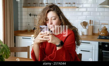 Eine Frau mit langen, welligen Haaren, die einen roten Pullover trägt, sitzt an einem Holztisch in einer modernen Küche und schaut aufmerksam auf ihr Smartphone Stockfoto