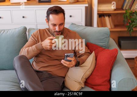 Blick aus einem hohen Winkel auf einen Mann im mittleren Erwachsenenalter, der Kaffee trank und E-Mails über das Telefon las, während er sich auf dem Sofa entspannte Stockfoto