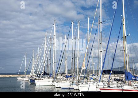 Weiße Segelboote, die an einem sonnigen Tag mit blauem Himmel und Wolken in einem Hafen vor Anker liegen, schaffen eine ruhige nautische Szene Stockfoto