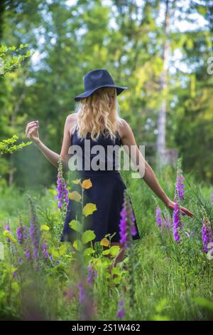 Junge blonde Frau mit schwarzem Kleid und Hut im Sommer mit Fuchshandschuhblumen im Waldportrait in der Natur. Junge blonde Frau mit schwarzem Kleid in Natur Stockfoto