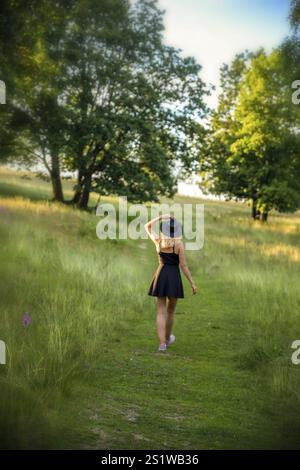 Junge blonde Frau, die an einem Sommerabend in der Natur spaziert und ein schwarzes Kleid trägt. Junge blonde Frau, die an einem Sommerabend in der Natur spaziert Stockfoto