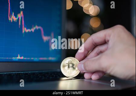 Eine Person hält eine Bitcoin-Münze vor einem Computerbildschirm. Auf dem Bildschirm wird ein Diagramm des Bitcoin-Preises angezeigt Stockfoto