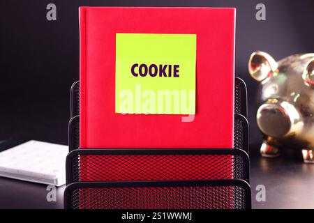 Ein leuchtend rotes Notizbuch zeigt eine Haftnotiz mit der Aufschrift COOKIE. Es befindet sich in einem Schreibtischordner neben einer glänzenden Schweinebank und schafft so ein verspieltes Werk Stockfoto