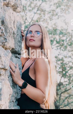 Frau, Steinmauer, Natur - Porträt einer Frau mit Brille und schwarzem Kleid, die sich draußen an eine Steinmauer lehnt. Stockfoto