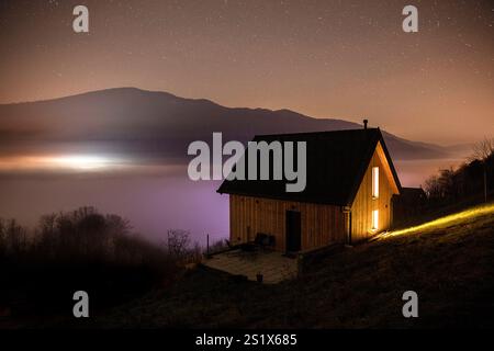 Schöne Holzhütte mit Lichtern in der Sternennacht, auf einem Hügel über dem Nebelmeer in der Weinlandschaft Dolenjske toplice, Slowenien Stockfoto