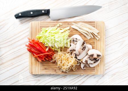 Gehacktes Gemüse auf einem hölzernen Schneidebrett mit einem Küchenmesser, Pilzen, Paprika, Sojasprossen und Kohl zum Kochen einer vegetarischen asiatischen Mahlzeit Stockfoto