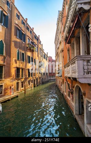 Ein schmaler venezianischer Kanal, gesäumt von farbenfrohen historischen Gebäuden mit Fensterläden und Balkonen, die das Sonnenlicht auf dem Wasser reflektieren. Ein paar s Stockfoto