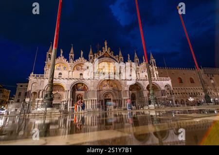 Beleuchtete Fassade des Markusdoms in Venedig, Italien, reflektiert auf nassem Pflaster unter einem dramatischen Abendhimmel und zeigt komplexe Architektur Stockfoto