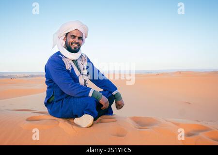 Ein Mann mittleren Alters posiert sitzend auf dem Wüstensand mit der Sahara im Hintergrund. Er trägt ein typisch marokkanisches Outfit. Djellaba, Turban und Stockfoto