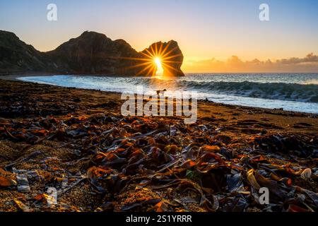 Durdle Door am zweiten Weihnachtsfeiertag 2022 spielt ein Hund in der Brandung, während die Wintersonne über Wolken am Horizont aufgeht und durch den natürlichen Felsbogen leuchtet Stockfoto