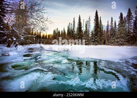 Eine ruhige Winterlandschaft mit einem teilweise gefrorenen Fluss umgeben von schneebedeckten Bäumen und einem klaren Himmel bei Sonnenuntergang. Stockfoto