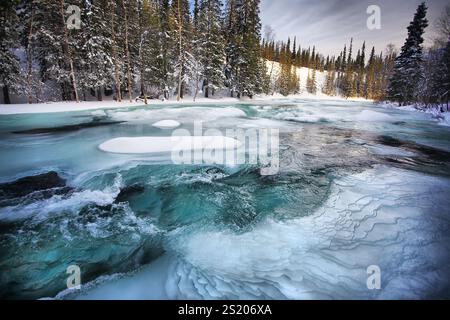 Ruhige Winterlandschaft mit teilweise gefrorenem Fluss, umgeben von schneebedeckten Bäumen. Eiswasser fließt langsam, kaltes Klima Stockfoto